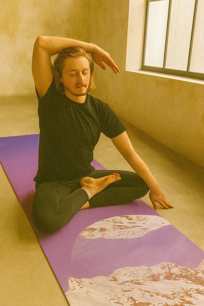 Man sitting on a purple yoga mat in a room with a window.