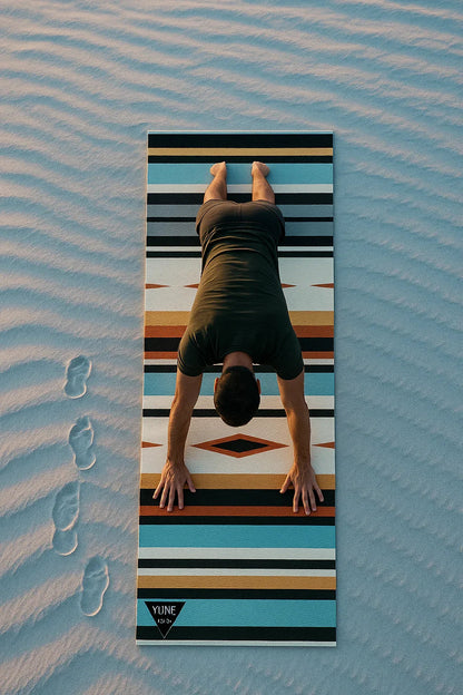 Person doing a handstand on a striped yoga mat with footprints in sand below.