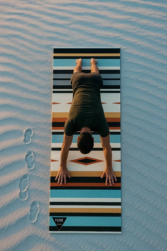 Person doing a handstand on a striped yoga mat with footprints in sand below.